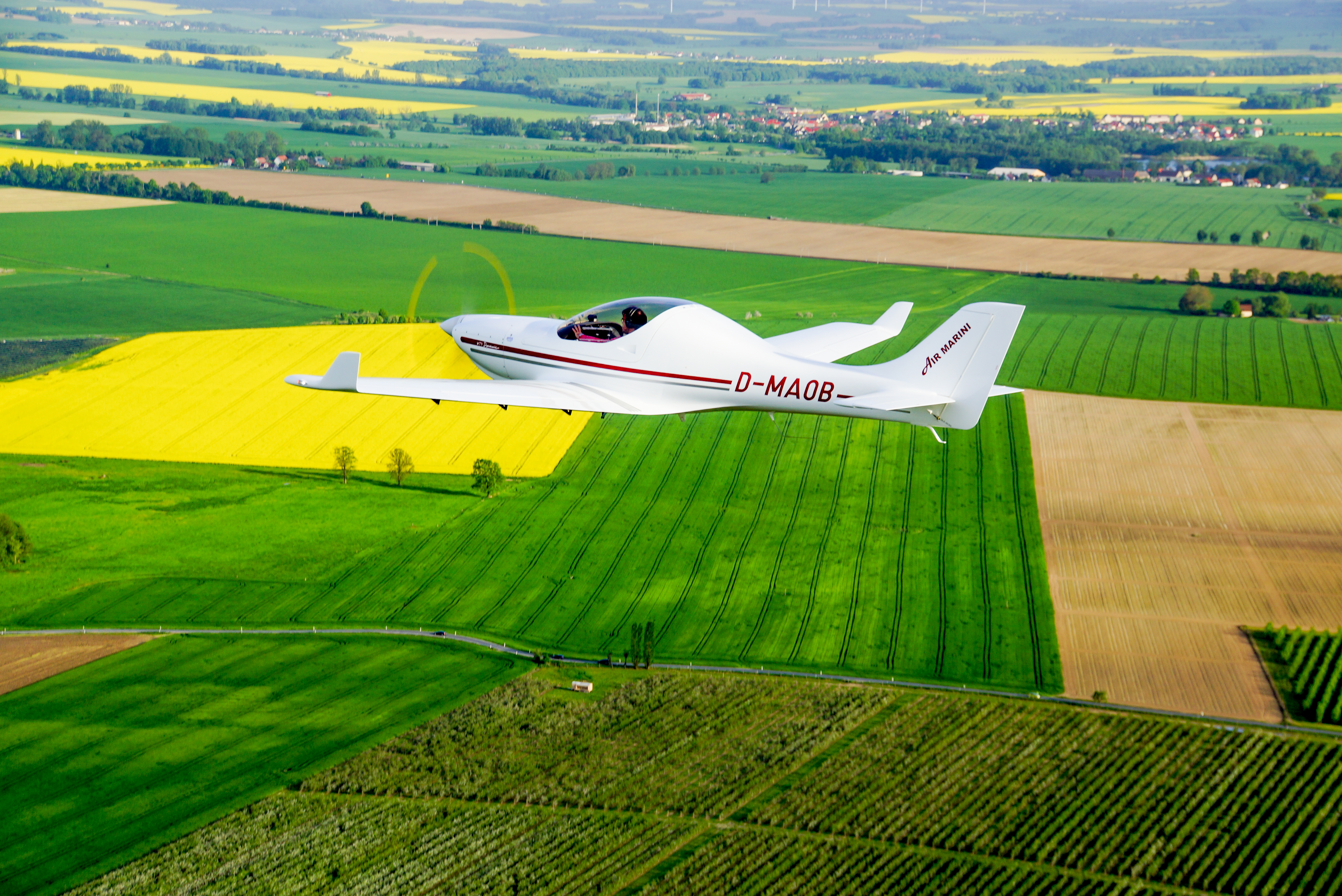 Ein weißes Leichtflugzeug mit der Aufschrift 'D-MA0B' fliegt über ein Mosaik aus grünen, gelben und braunen Feldern.