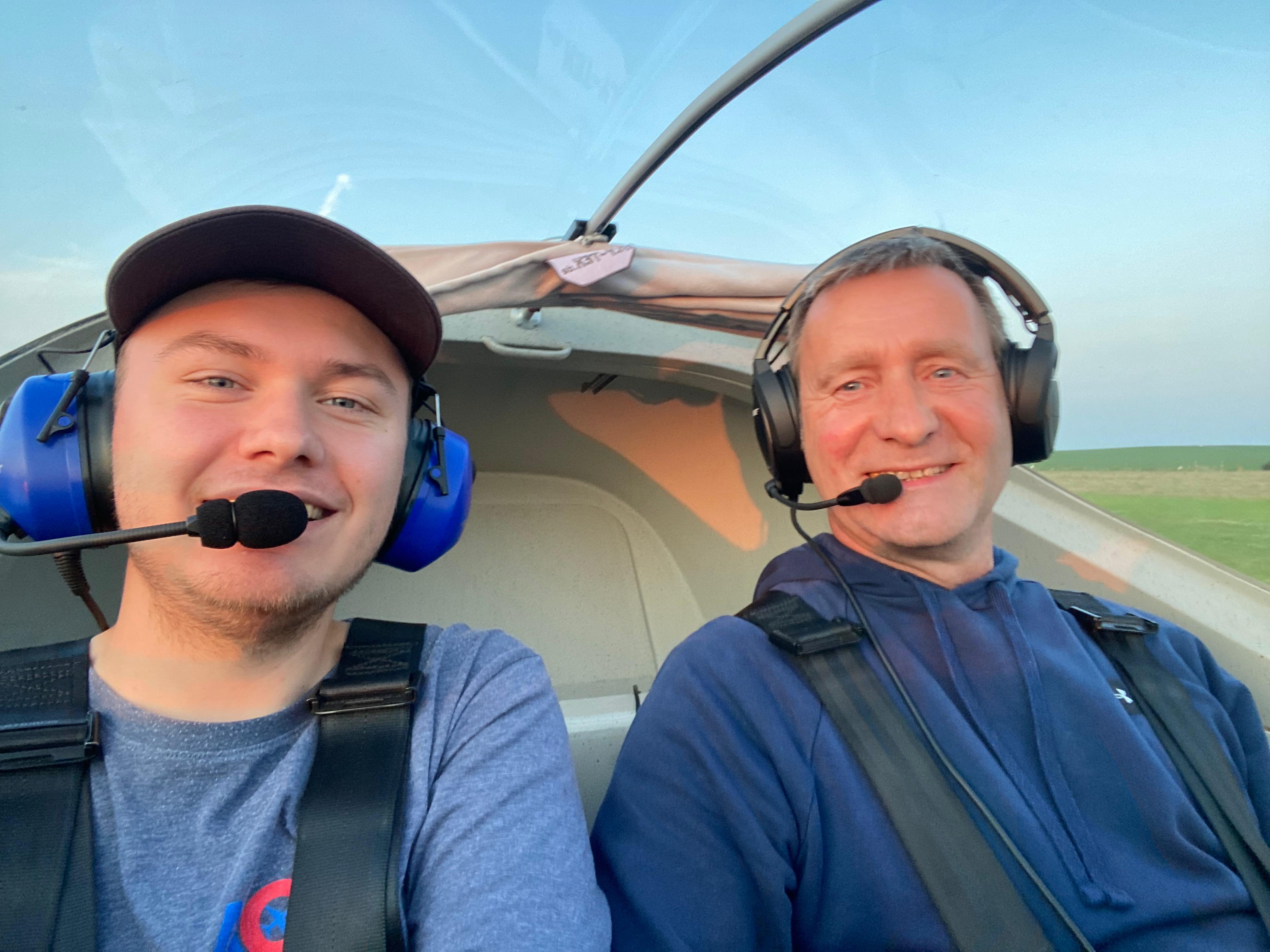 Zwei Männer im Cockpit eines Flugzeugs mit Kopfhörern, die ein Selfie machen. Es ist sonnig und im Hintergrund ist der Himmel blau.