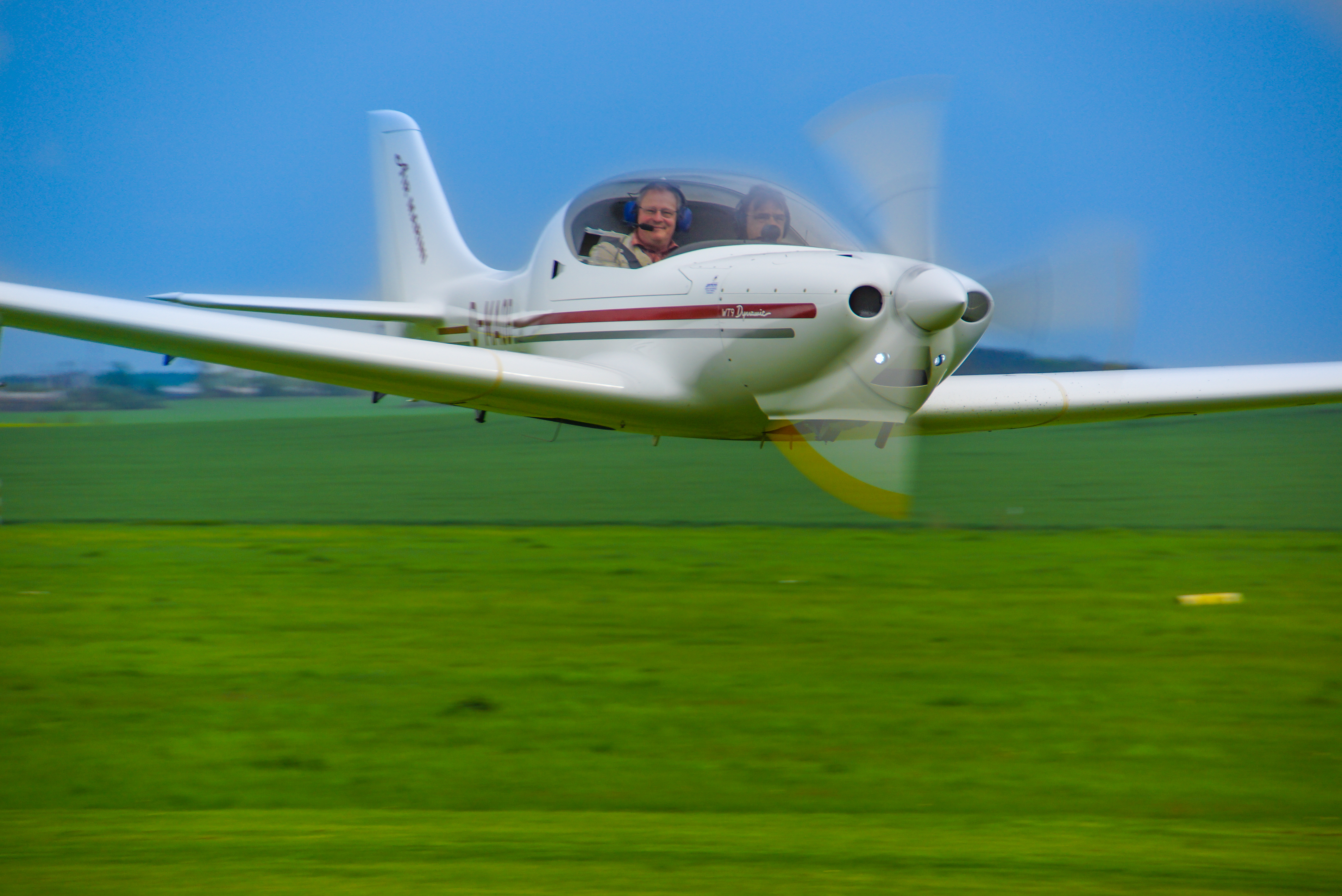 Weißes Leichtflugzeug fliegt knapp über einem grünen Feld bei blauem Himmel