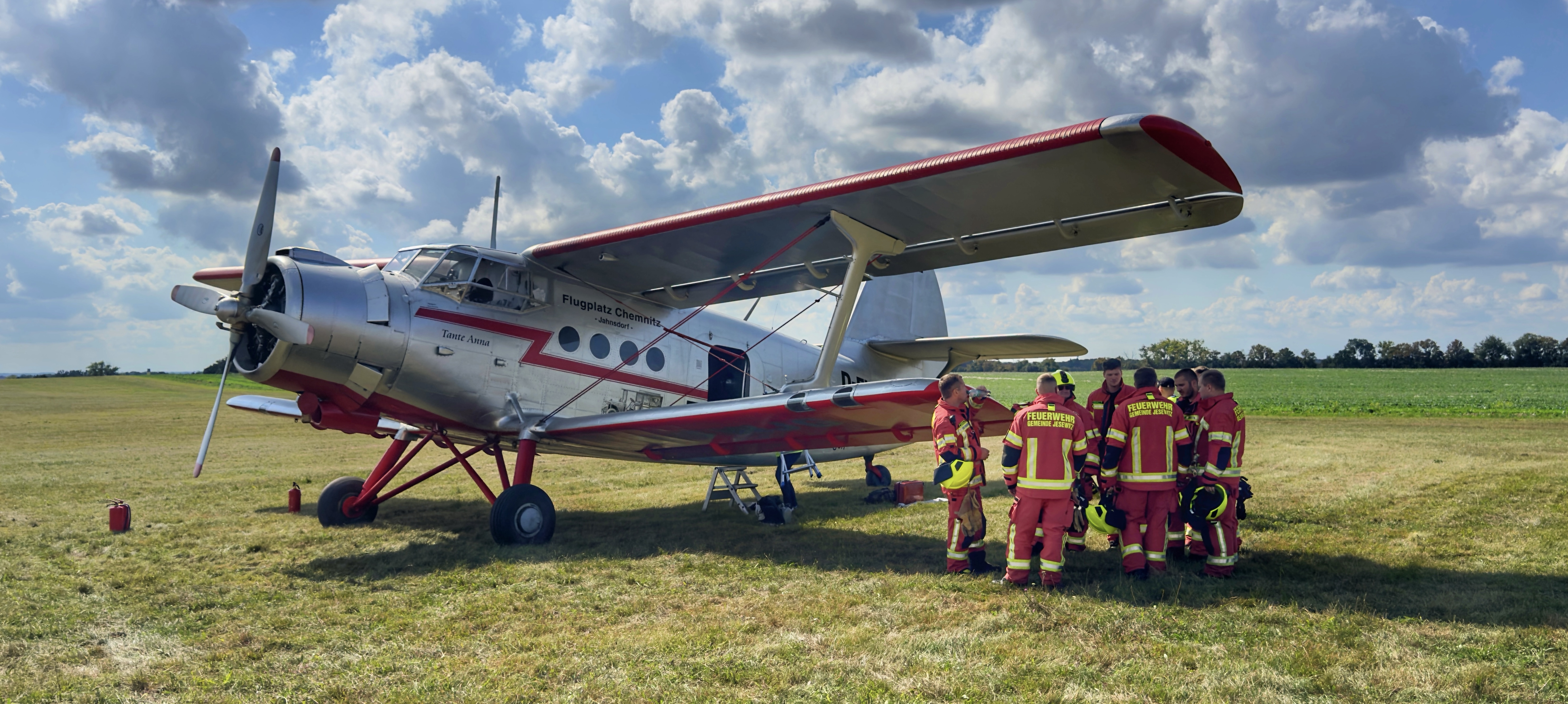 Rundflugtage mit der Antonov AN-2 auf dem Flugplatz Taucha – 03. und 04. Oktober 2025 Silbernes Flugzeug mit roter Markierung auf einer Wiese, umgeben von einer Gruppe Feuerwehrleute in Einsatzkleidung, unter blauem Himmel mit Wolken