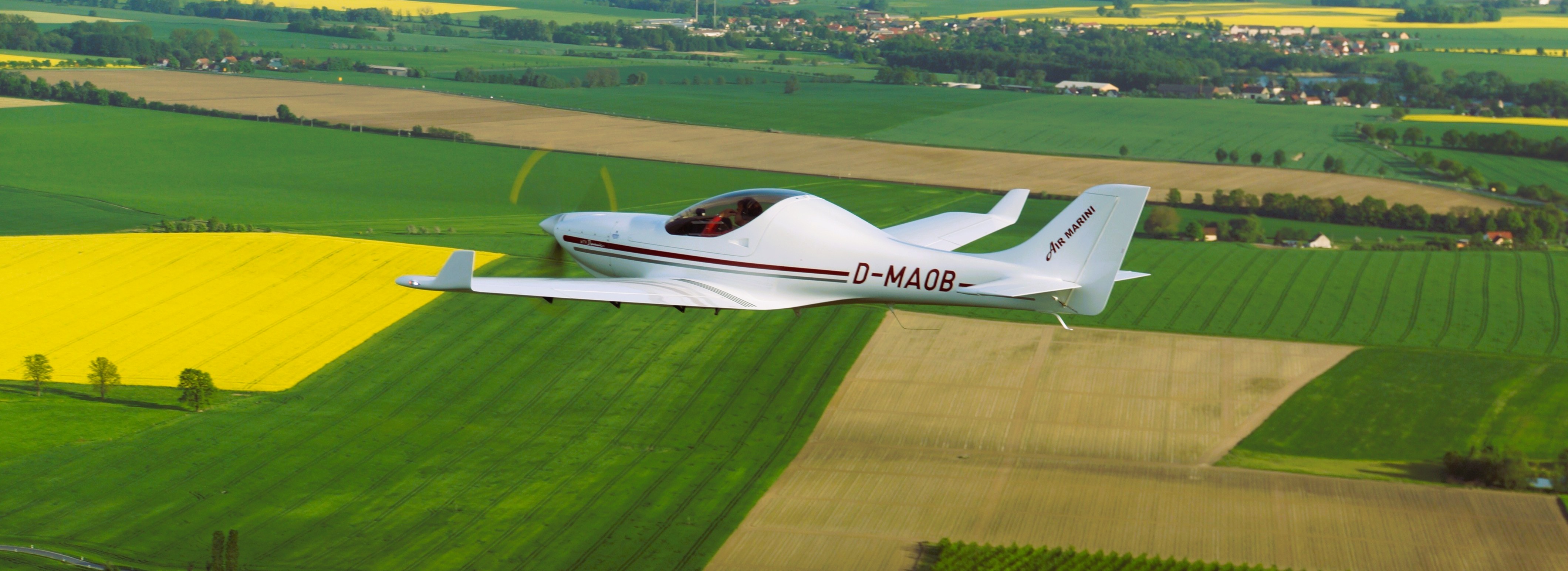 Ein weißes Leichtflugzeug fliegt über weitläufige Felder, die in verschiedenen Grüntönen und leuchtendem Gelb erstrahlen.