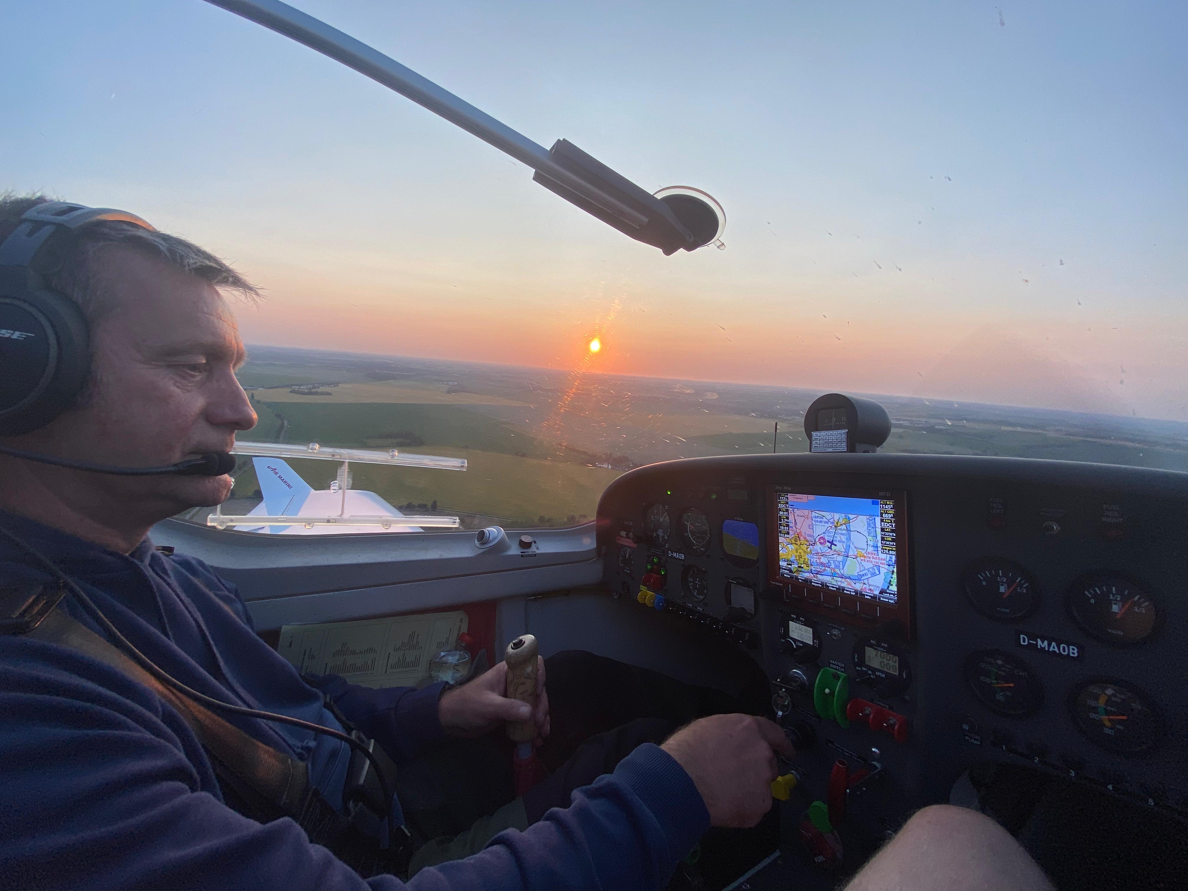 Ein Pilot steuert ein Leichtflugzeug im Sonnenuntergang. Das Cockpit zeigt diverse Instrumente und ein Display mit Flugdaten.