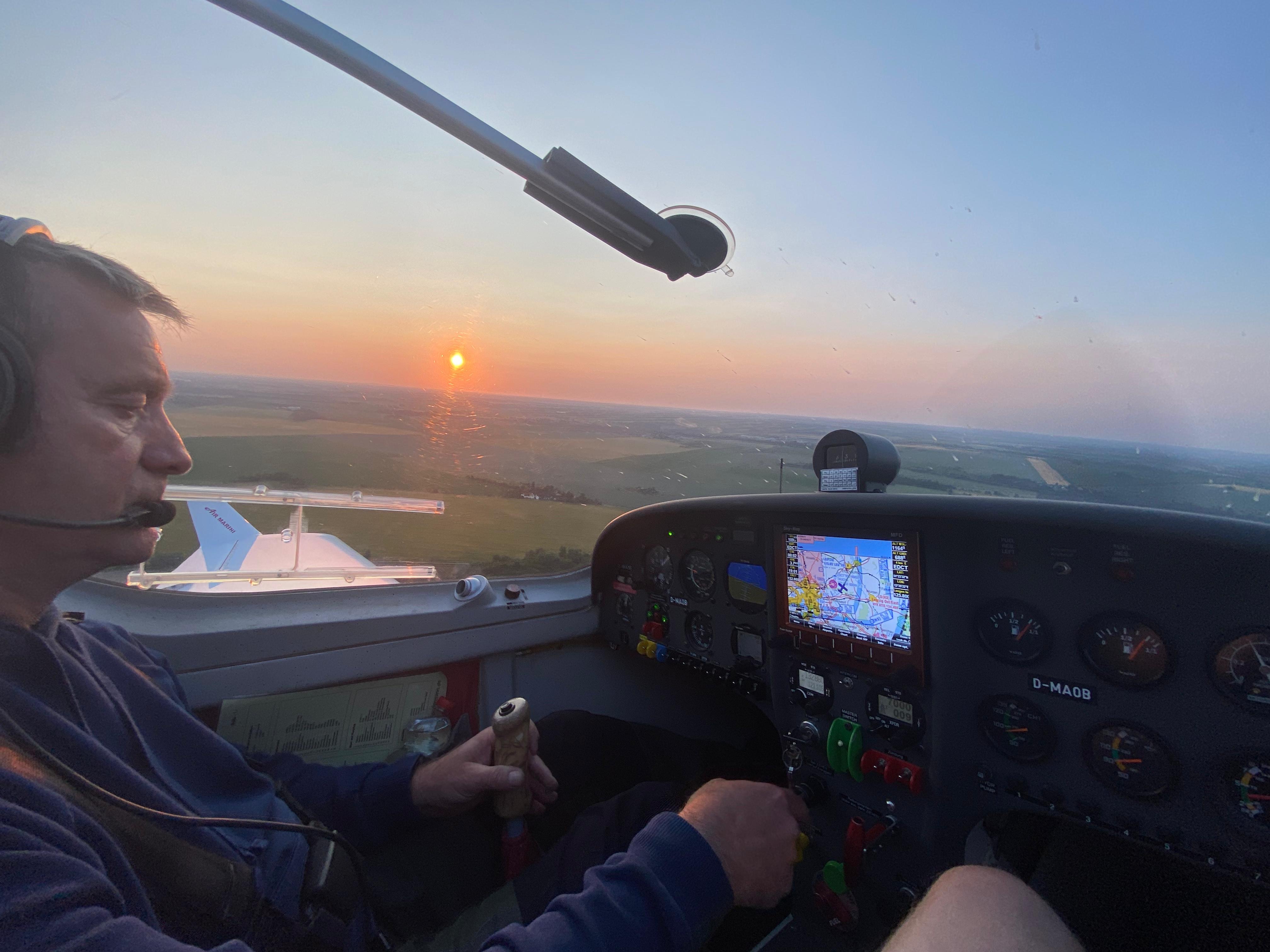 Ein Pilot im Cockpit eines Flugzeugs steuert während eines Sonnenuntergangs über einer Landschaft. Das Armaturenbrett mit Instrumenten ist sichtbar.