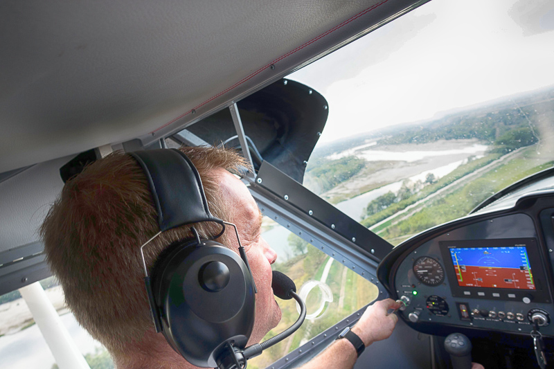 Nahaufnahme eines Piloten im Cockpit eines Kleinflugzeugs, der über eine grüne Flusslandschaft fliegt