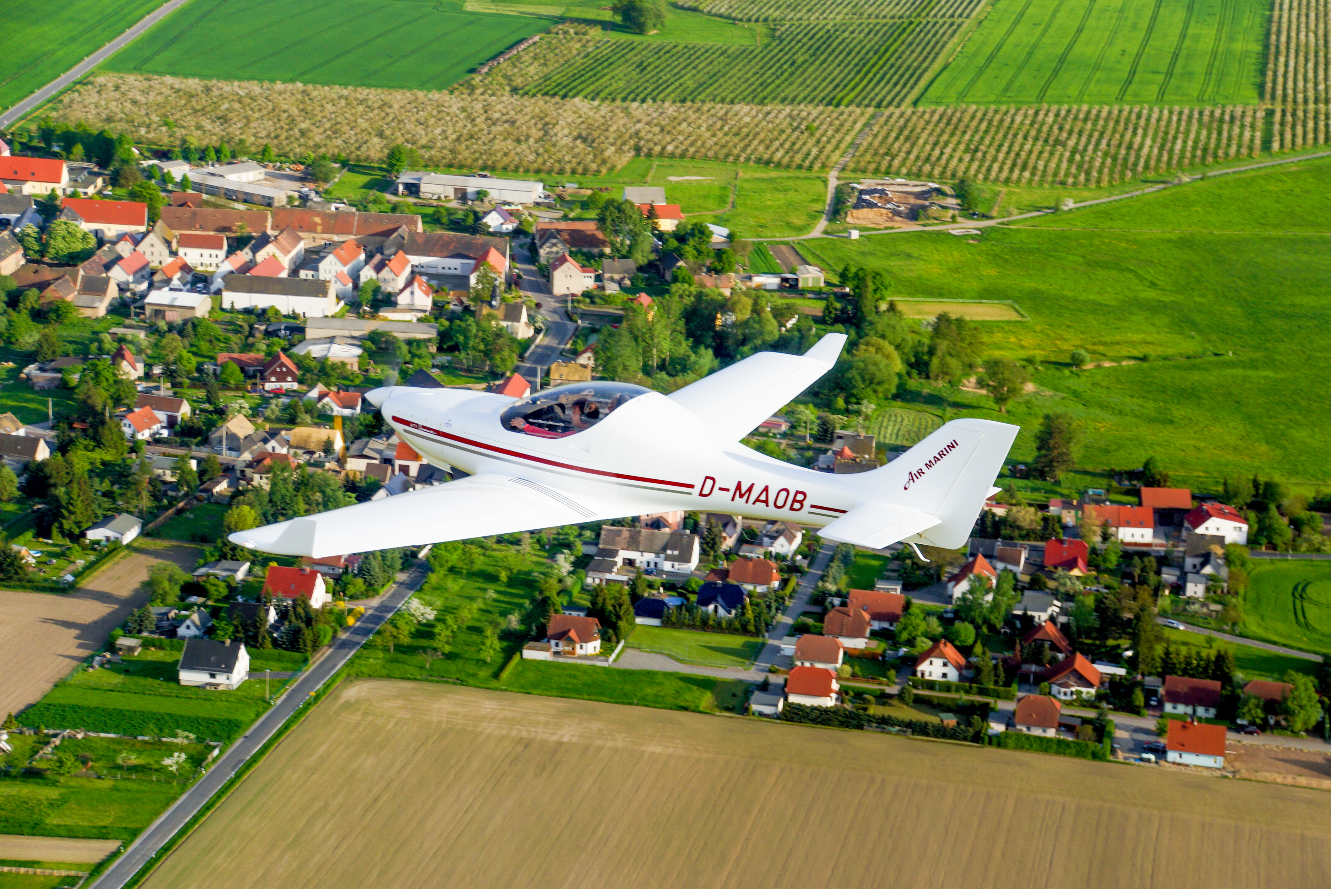 Ein weißes Leichtflugzeug fliegt über eine grüne ländliche Region in Deutschland, mit kleinen Häusern und Feldern sichtbar.