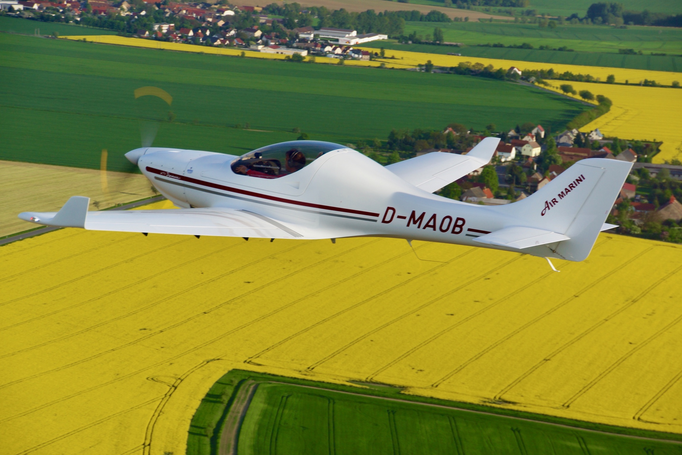 Weißes Sportflugzeug fliegt über ein gelbes Rapsfeld, im Hintergrund eine ländliche Landschaft und ein Dorf