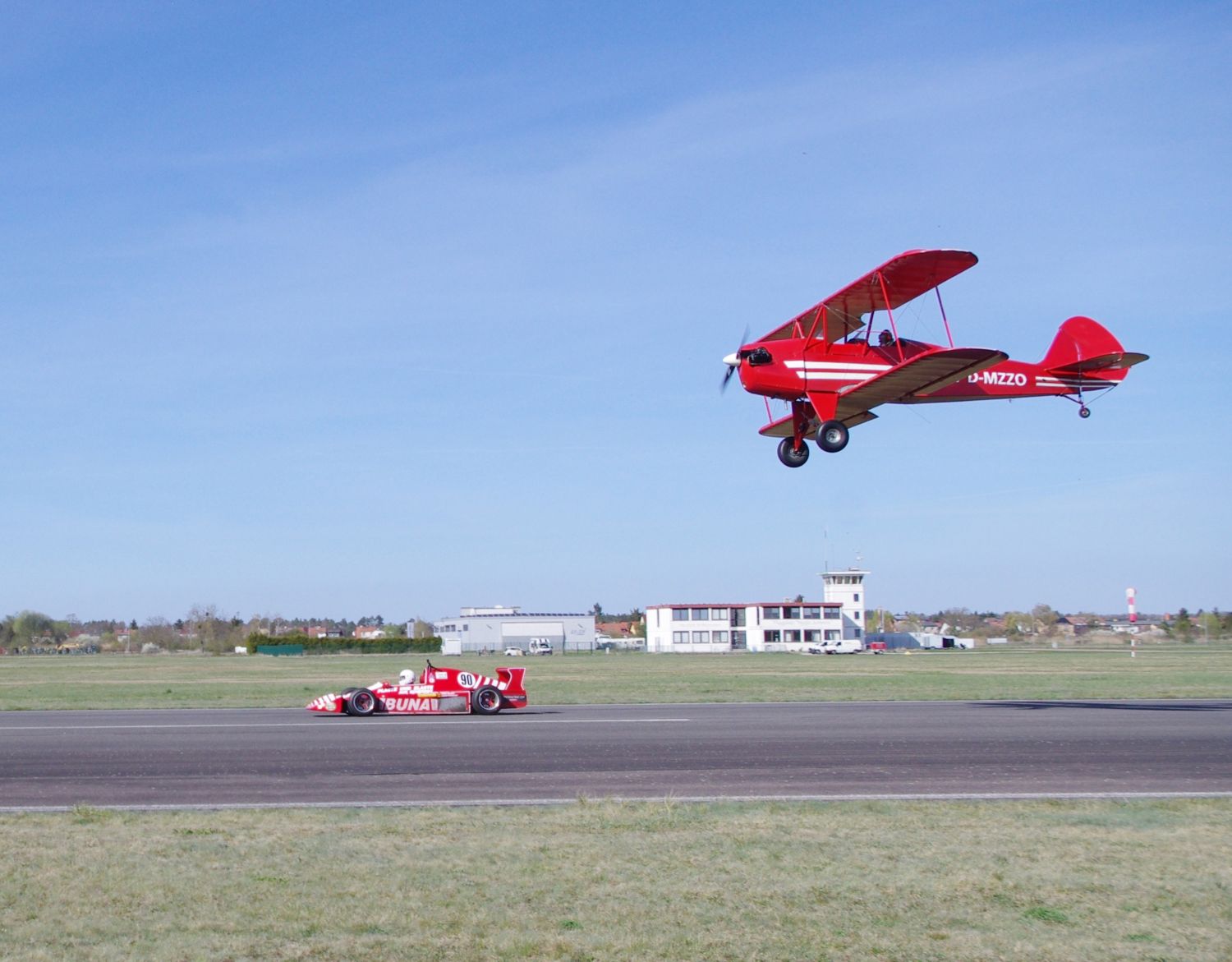 Ein rotes Oldtimer-Flugzeug fliegt über einen roten Rennwagen auf einer Start- und Landebahn.