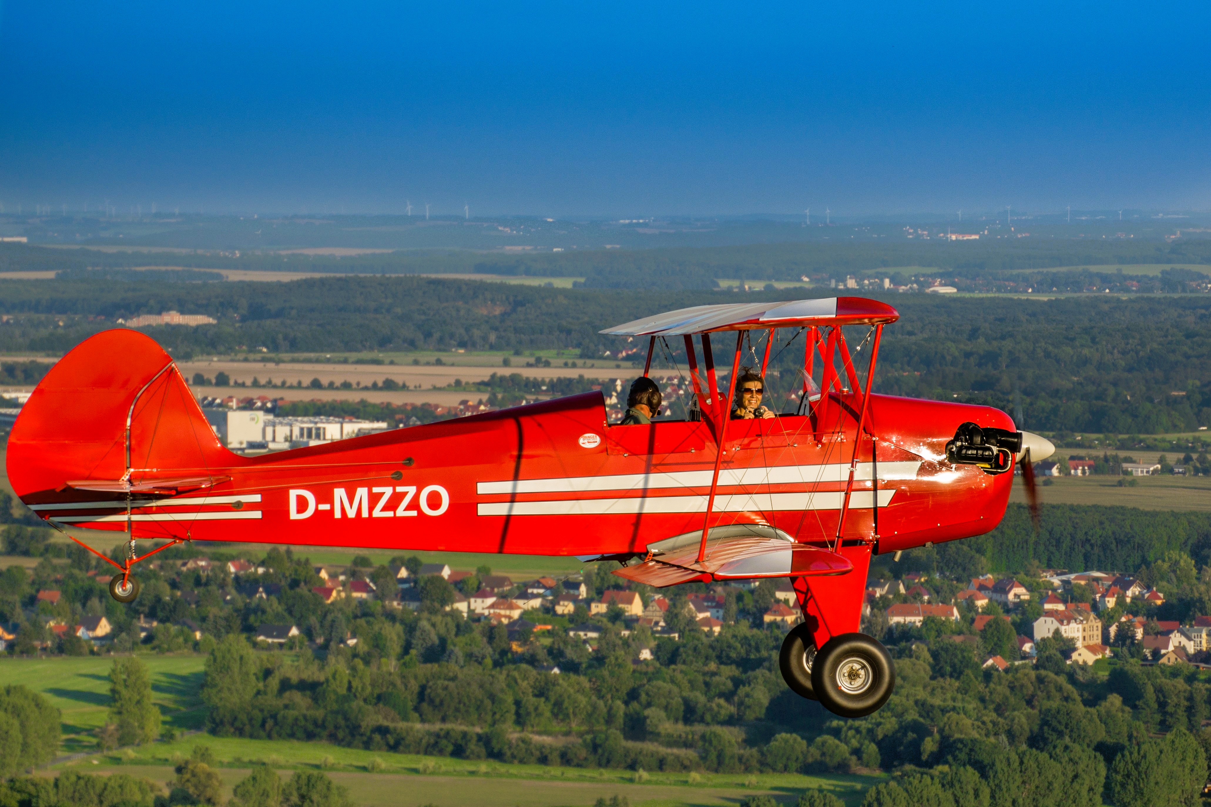 Rotes Doppeldeckerflugzeug mit der Kennung D-MZZO fliegt über eine grüne Landschaft mit Feldern und Häusern im Hintergrund.